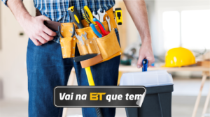 Cinto de couro preso à cintura de um homem, contendo diversas ferramentas. Martelo, chave de fenda, alicate, fita métrica, furadeira, nível, serra, chave de boca ou chave inglesa. Na mão o homem carrega uma caixa de ferramentas e ao fundo é possível ver alguns equipamentos de proteção individuais (EPIs).
